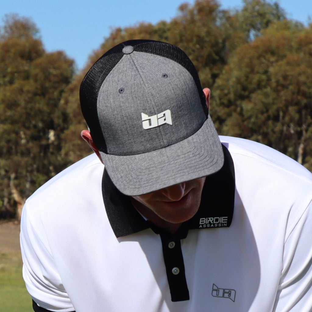 Person wearing a Tour golf cap from Birdie Assassin with a logo, standing outdoors with trees in the background.