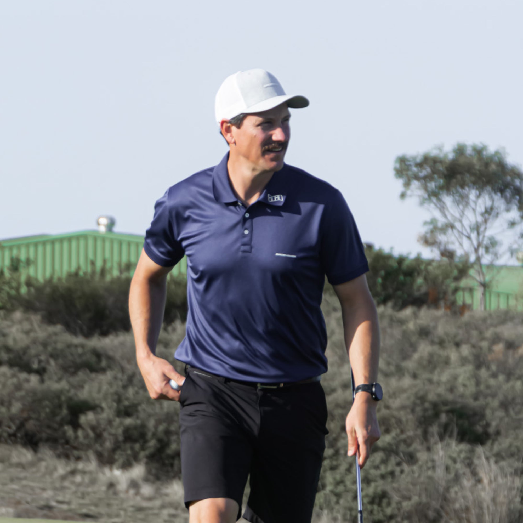 Man in a Bourne golf polo from Birdie Assassin and white cap holding a golf club on a golf course.
