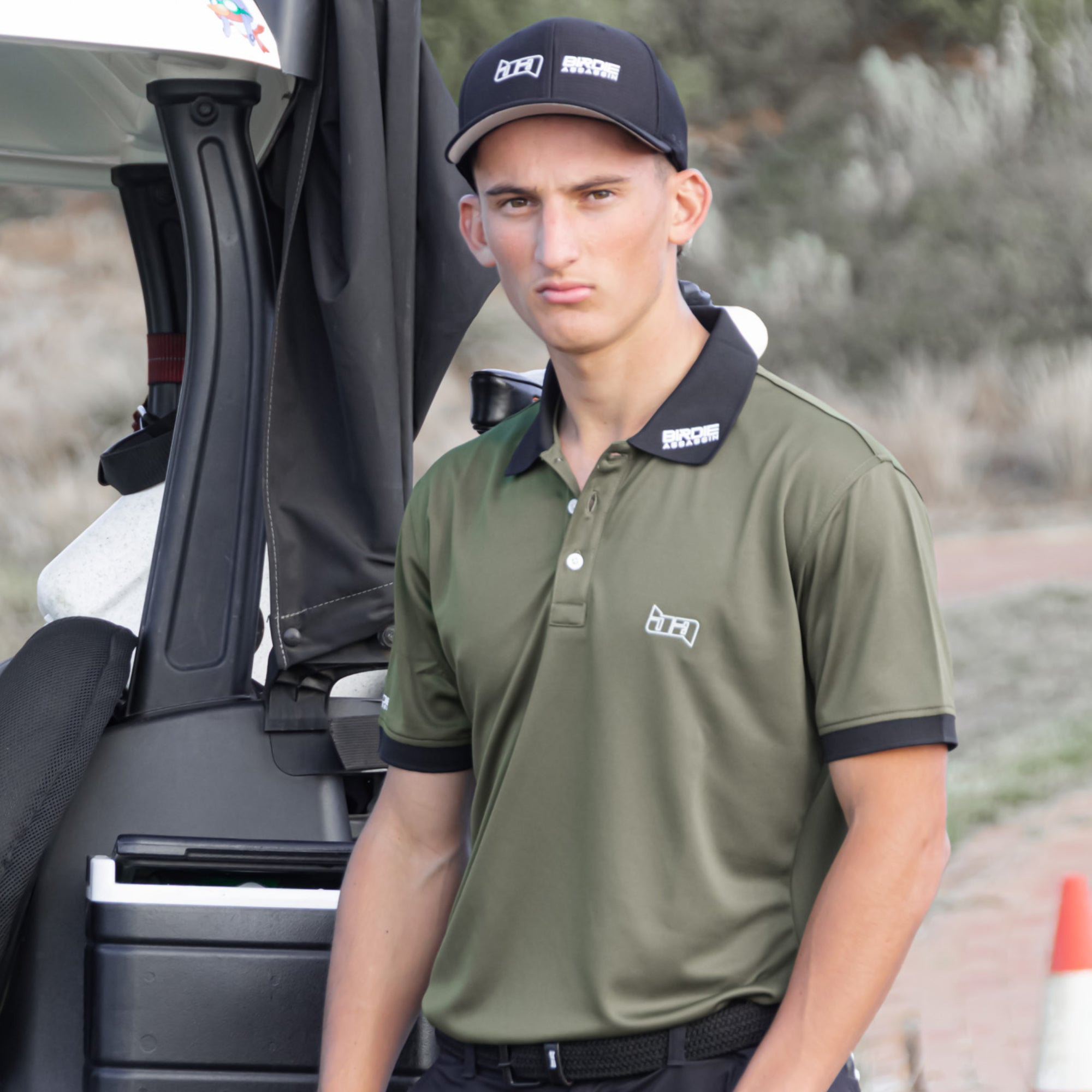Man wearing a Marksman golf polo from Birdie Assassin standing next to a golf cart.