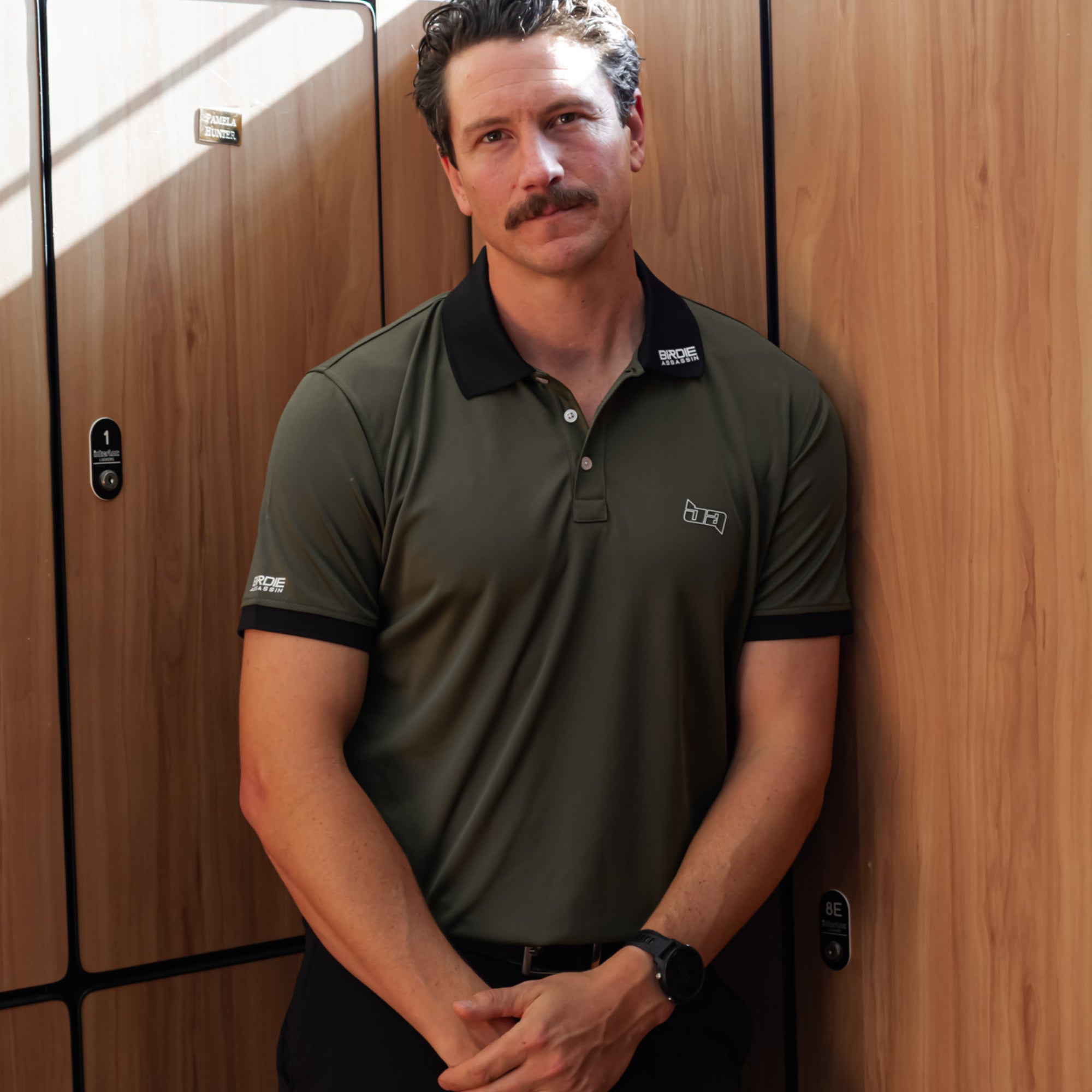 Man wearing a Marksman golf polo from Birdie Assassin against a wooden locker room.